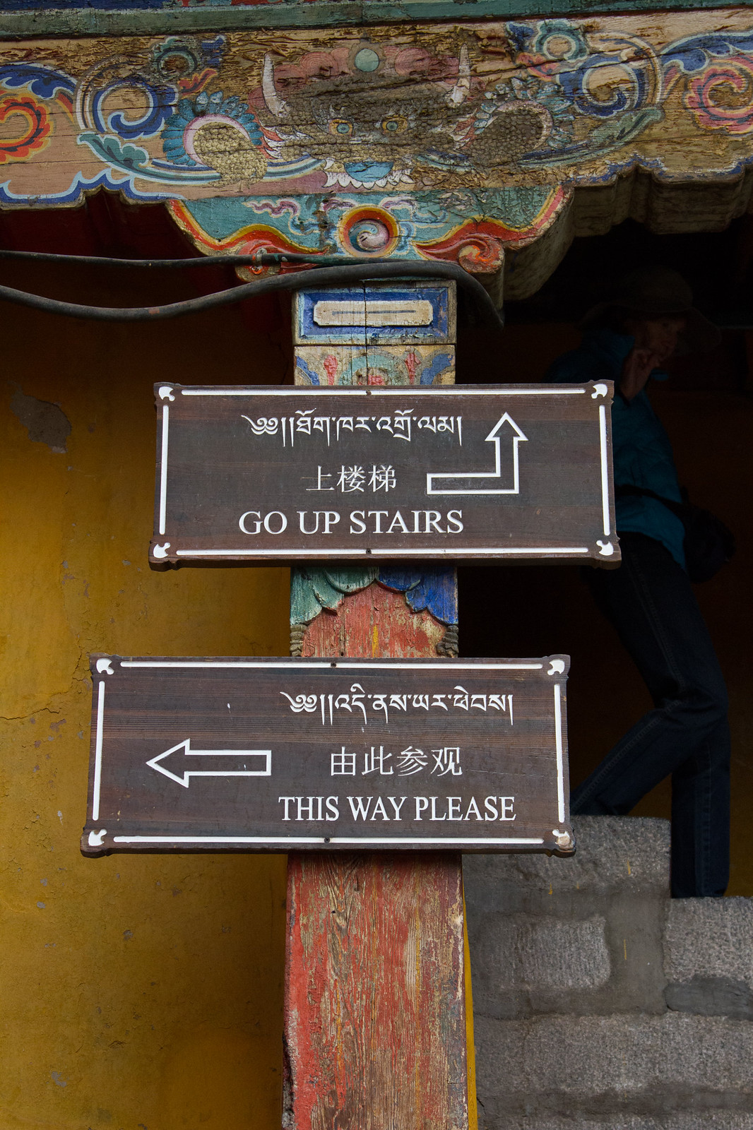 Trilingual monastery signs reading "GO UP STAIRS" and "THIS WAY PLEASE" below an ornate painted eave.