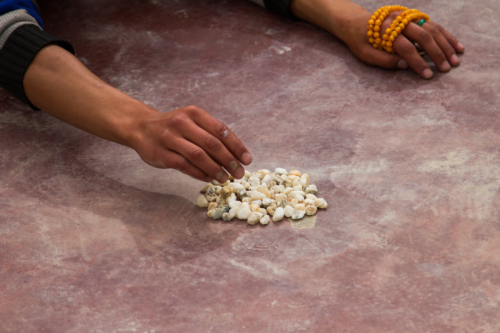 Hands reaching toward a pile of small white pebbles on stone, one wrist wrapped in amber prayer beads.