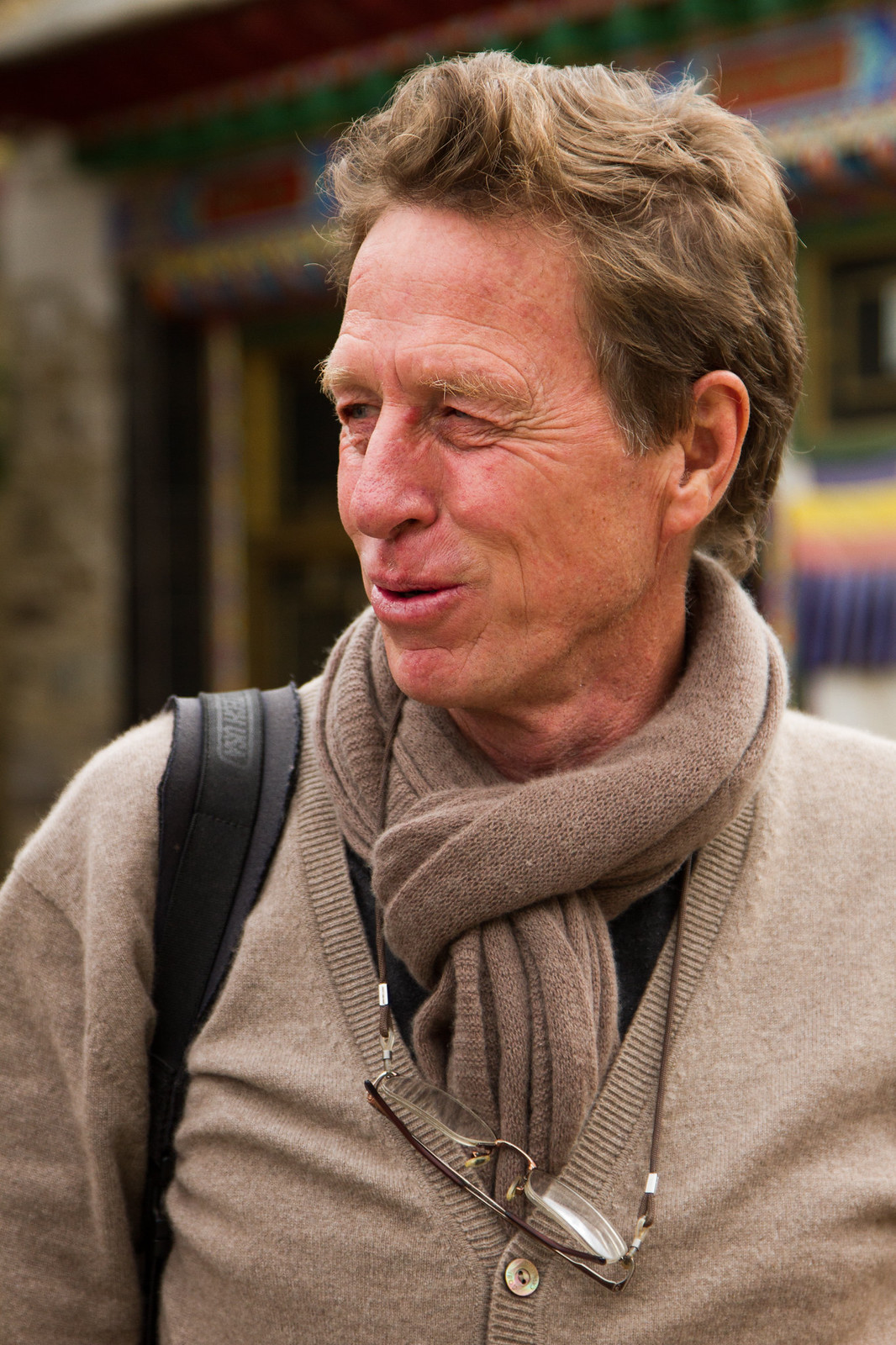 An older man in a tan sweater and scarf, glasses hanging from his neck, before a colorful Tibetan building.