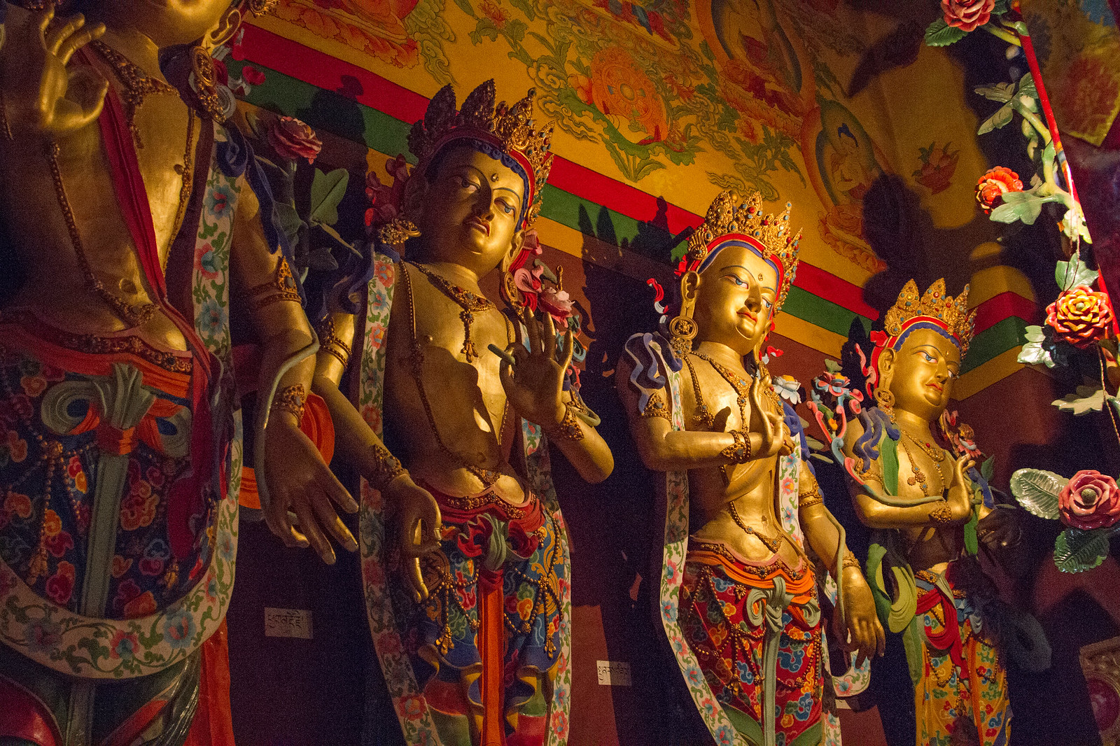 Row of gilded, jewel-crowned bodhisattva statues beneath a painted ceiling in a Tibetan monastery.