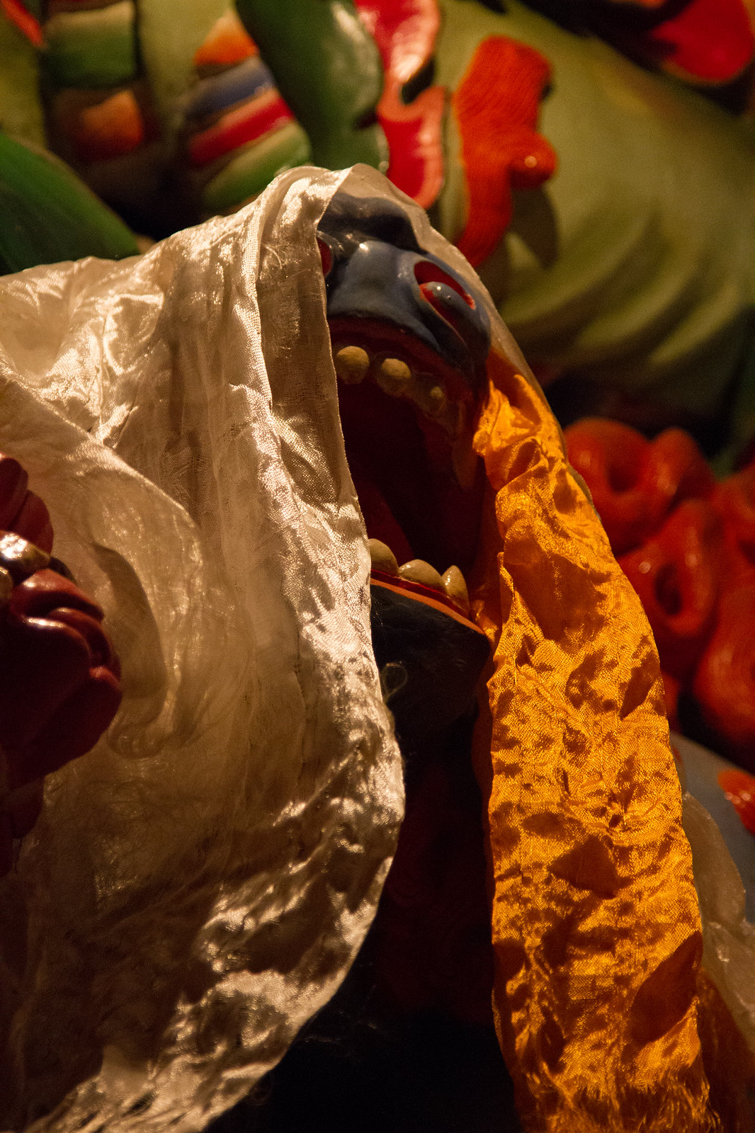 A colorful wrathful Tibetan deity statue with fanged open mouth, draped in white and gold silk offering scarves.