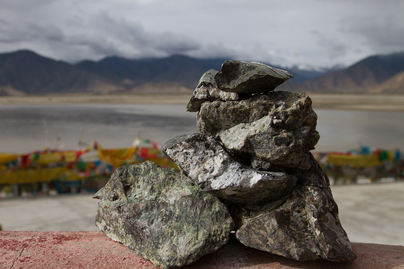 A cairn of stacked rocks on a red ledge with Tibetan prayer flags, a lake, and mountains behind.