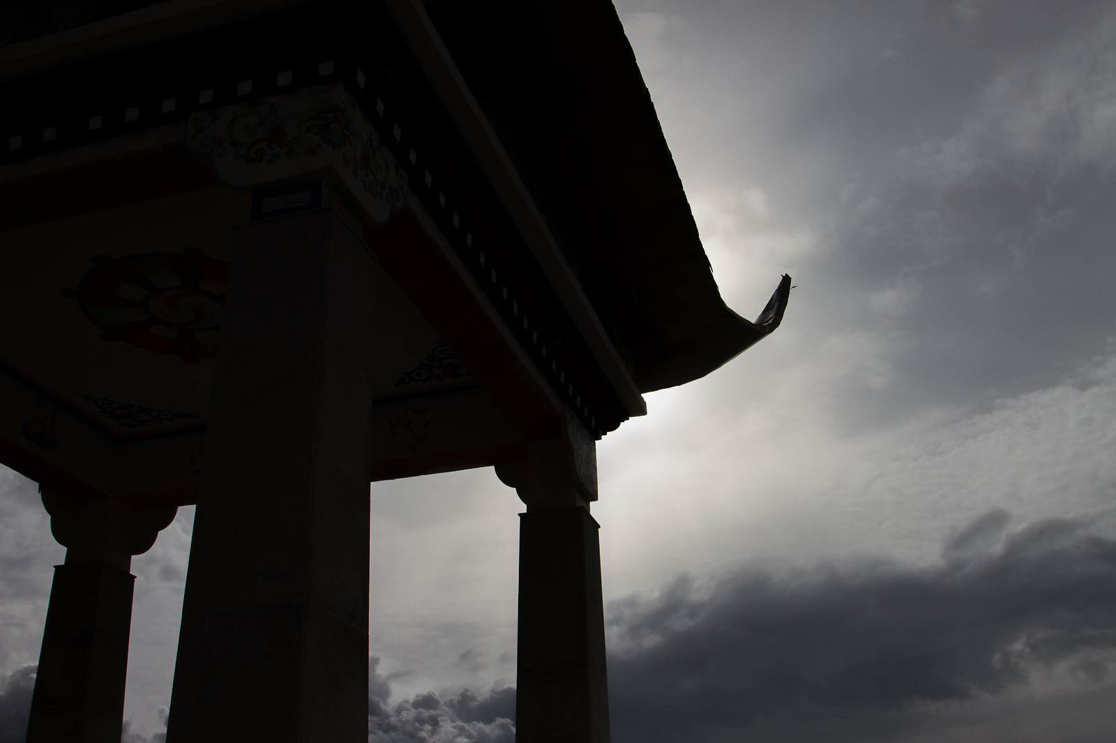 The upswept eaves and painted dharma-wheel of a Tibetan temple gateway silhouetted against a brooding grey sky.
