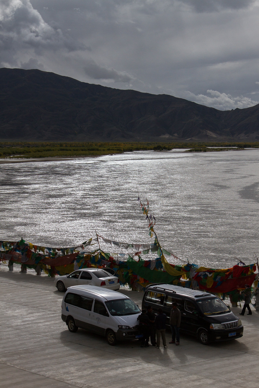 Colorful Tibetan prayer flags strung along a shimmering river with vans, cars, and people parked below dark mountains.