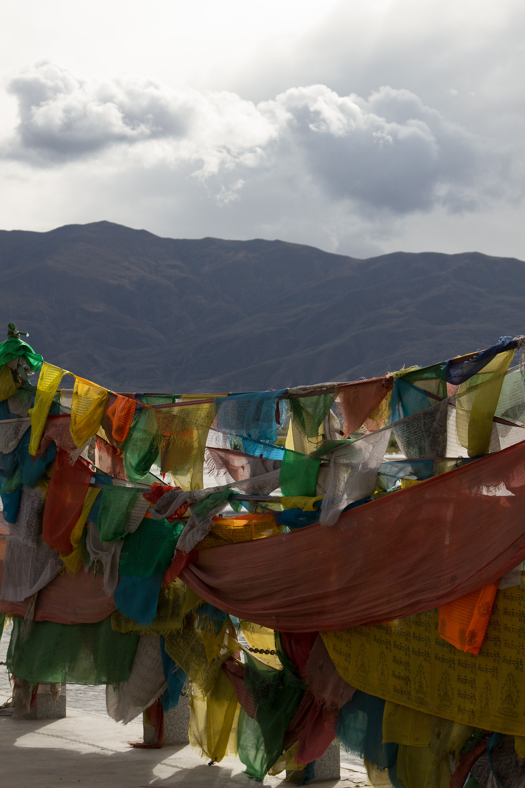 Colorful Tibetan prayer flags strung across a rooftop against a dark mountain ridge and cloudy sky.