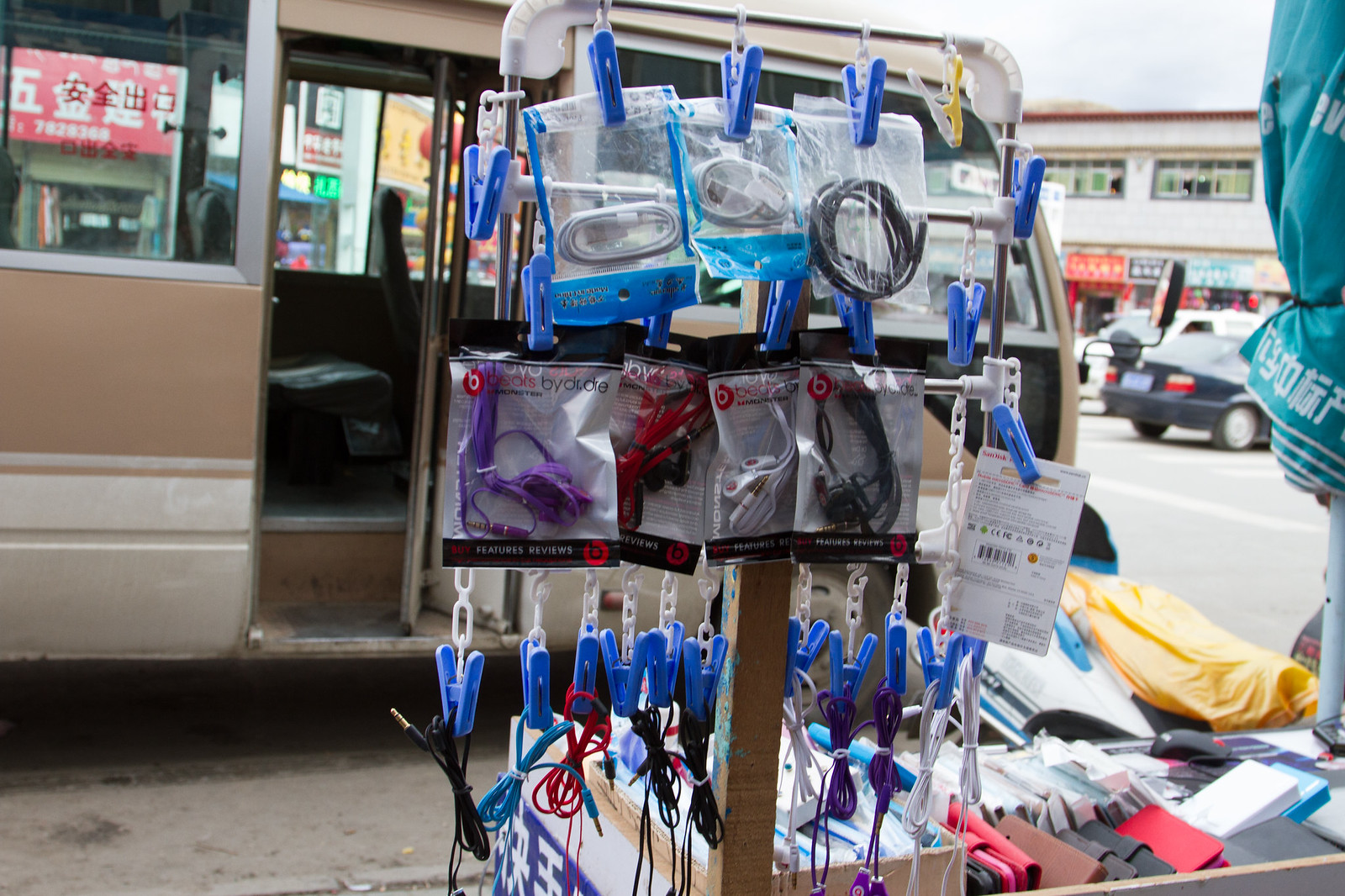 Street vendor stand selling knockoff Beats by Dr. Dre earphones and cables clipped to a rack beside a bus.