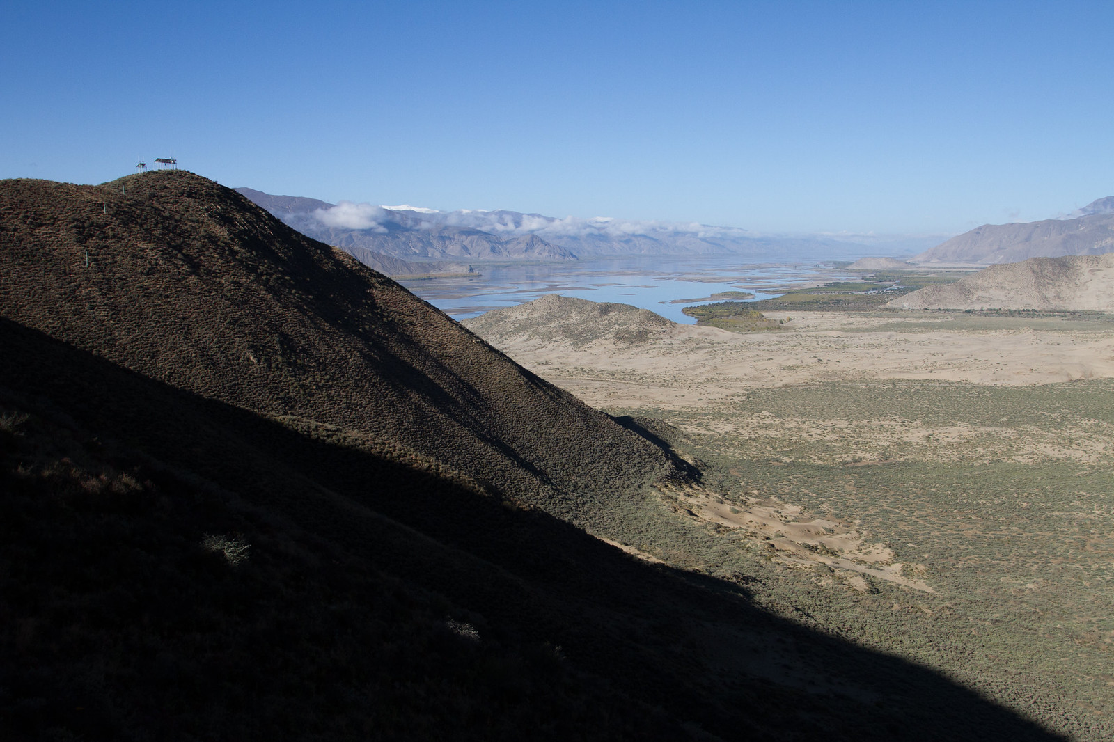 Prayer flags crown a shadowed ridge above a broad Tibetan river valley with braided water and distant snow-dusted peaks.