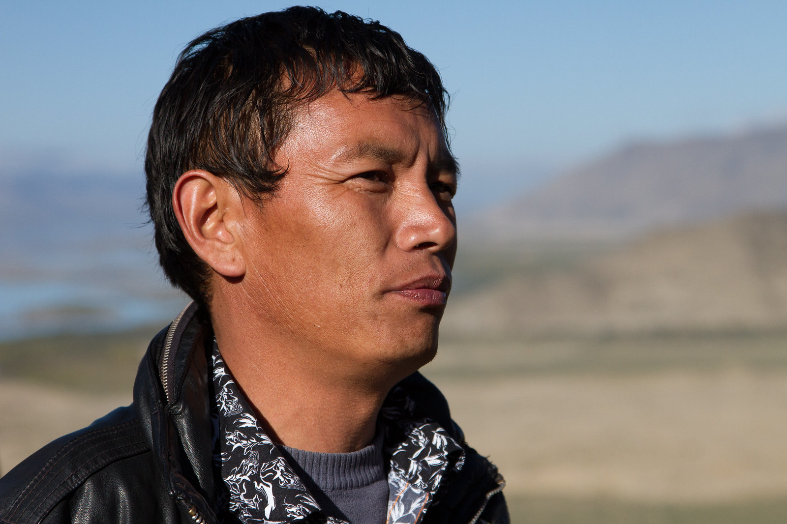 Weathered man in a leather jacket and floral scarf gazes into the distance across a sunlit Tibetan plateau.