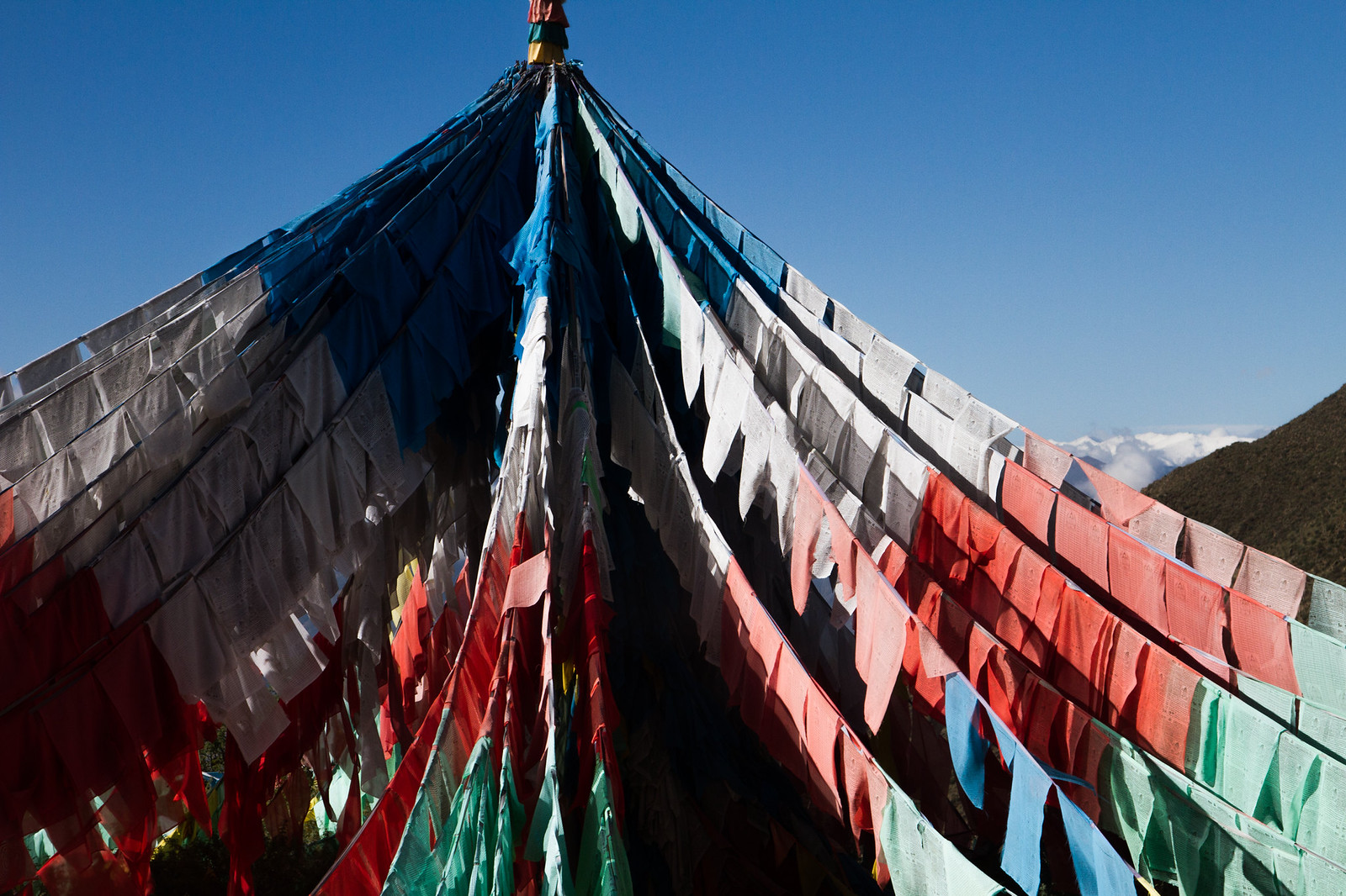 Rows of colorful Tibetan prayer flags strung from a high point against a deep blue sky, mountains beyond.