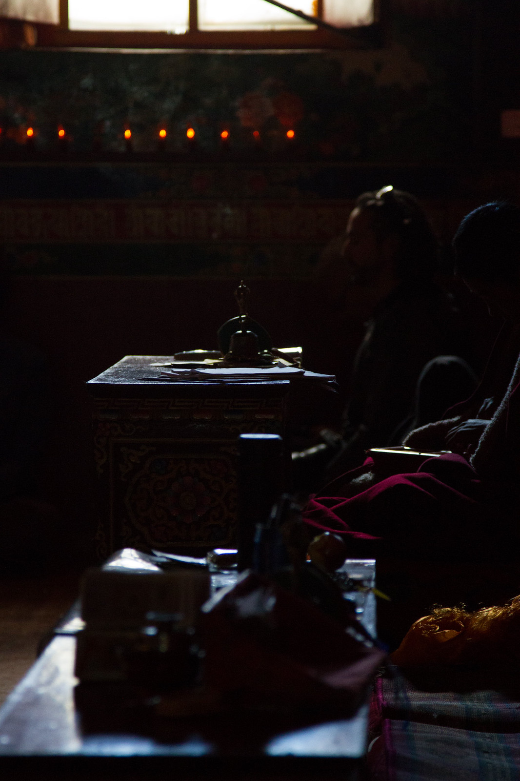 Butter lamps glow along a dim monastery wall as monks in maroon robes sit beside an ornate altar table.