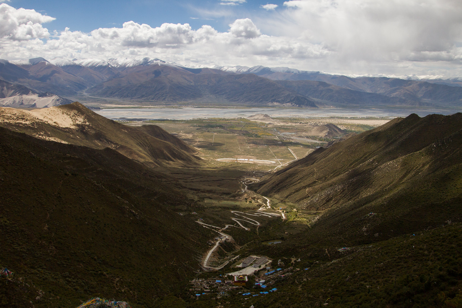 A winding dirt road descends through green Tibetan mountain valleys toward a small village and broad river plain.