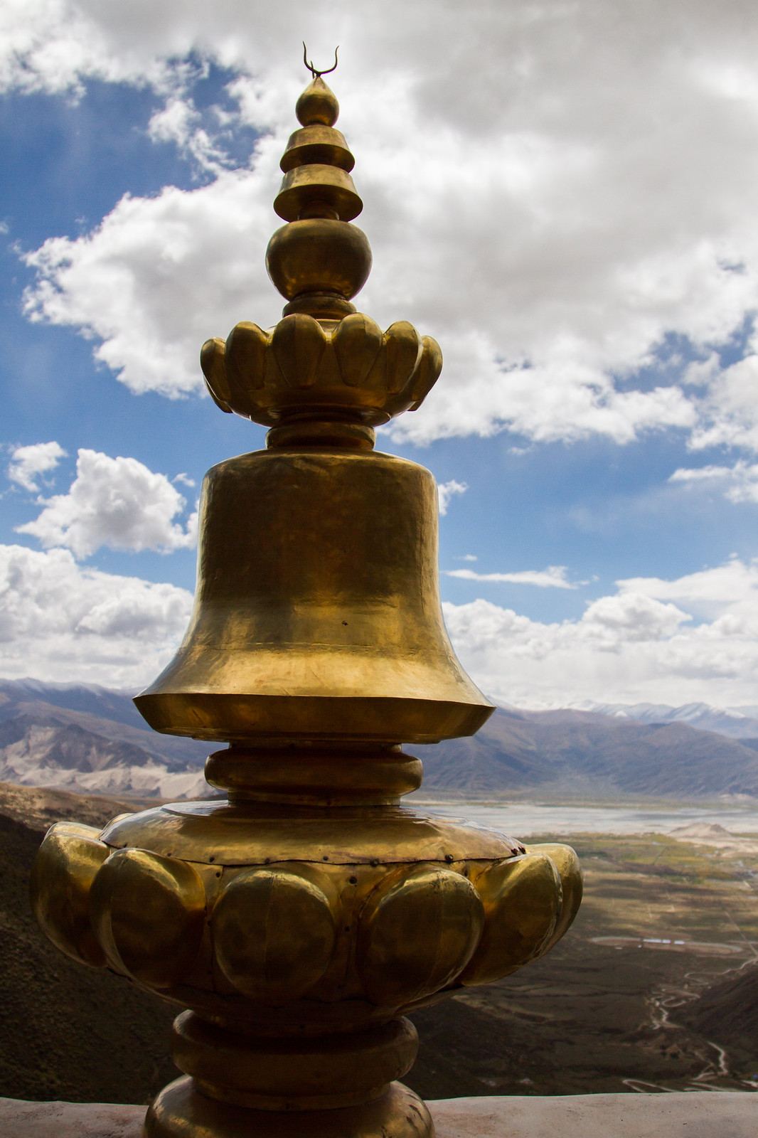 Golden Buddhist stupa ornament atop a monastery roof overlooking a broad Tibetan valley and mountains under drifting clouds.
