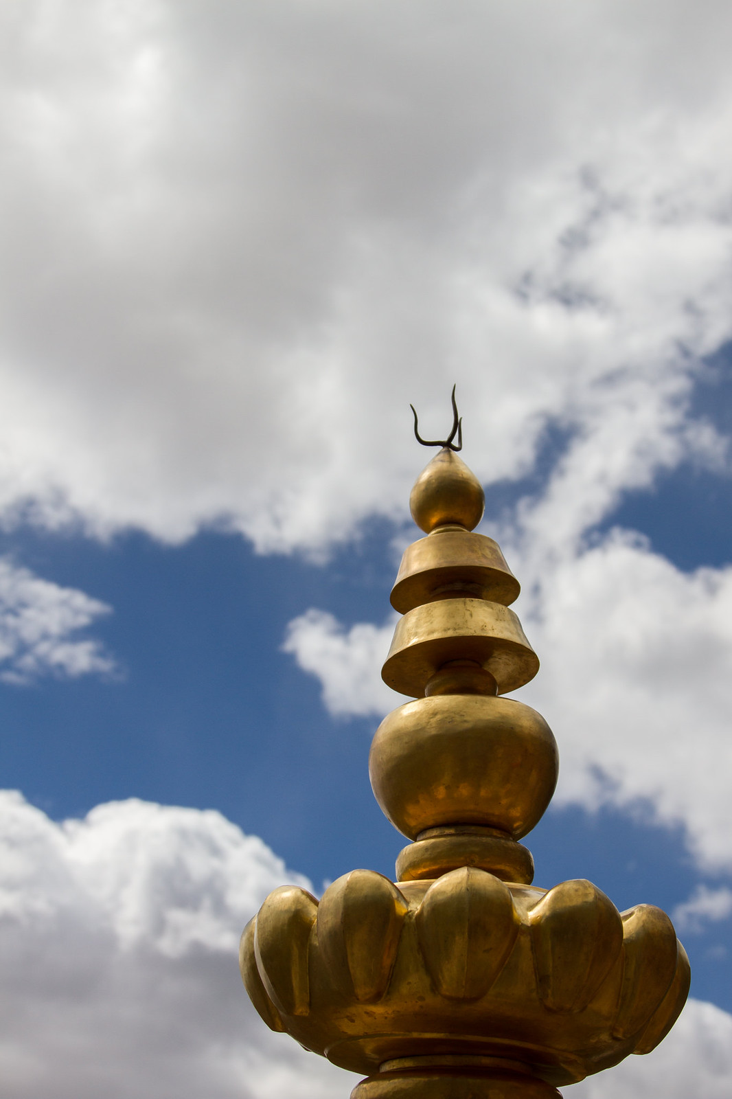 A golden Tibetan Buddhist rooftop finial with tiered lotus and orb shapes against a bright cloudy blue sky.