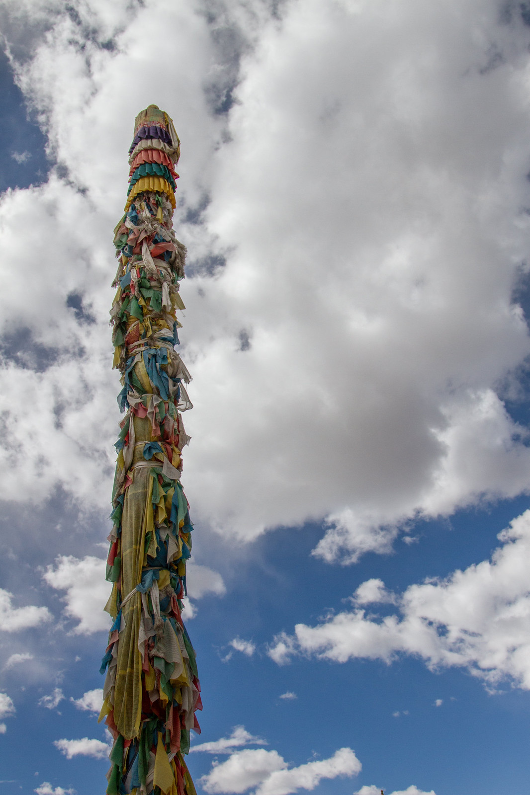 A tall Tibetan prayer pole wrapped in layers of colorful weathered prayer flags against a cloudy blue sky.