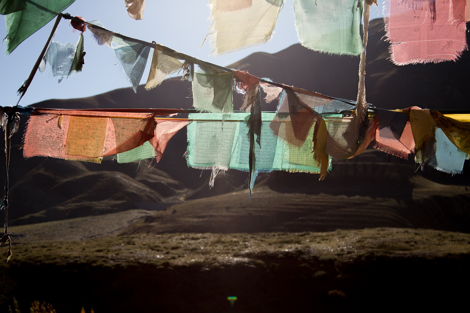 Weathered Tibetan prayer flags strung across a barren mountain valley, backlit by low sun.