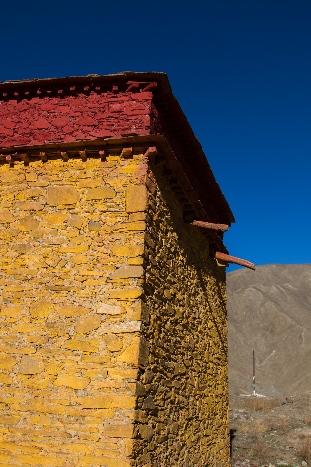 Sunlit corner of a Tibetan stone building with yellow-ochre walls and a red-painted upper band, against a deep blue sky and barren mountain.