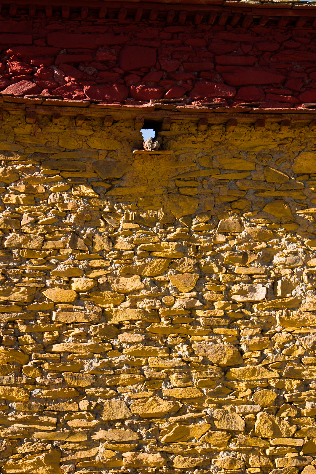 Sunlit Tibetan stone wall with a rough-hewn masonry base and a rust-red painted upper band.