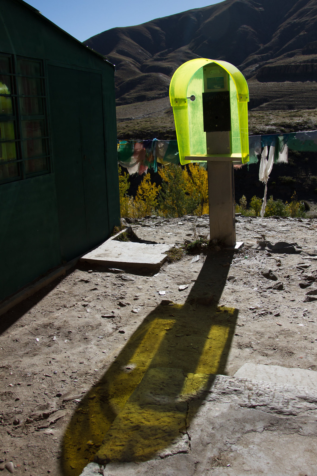 A neon-yellow phone booth casts a green shadow beside a green hut, with prayer flags and barren mountains behind.