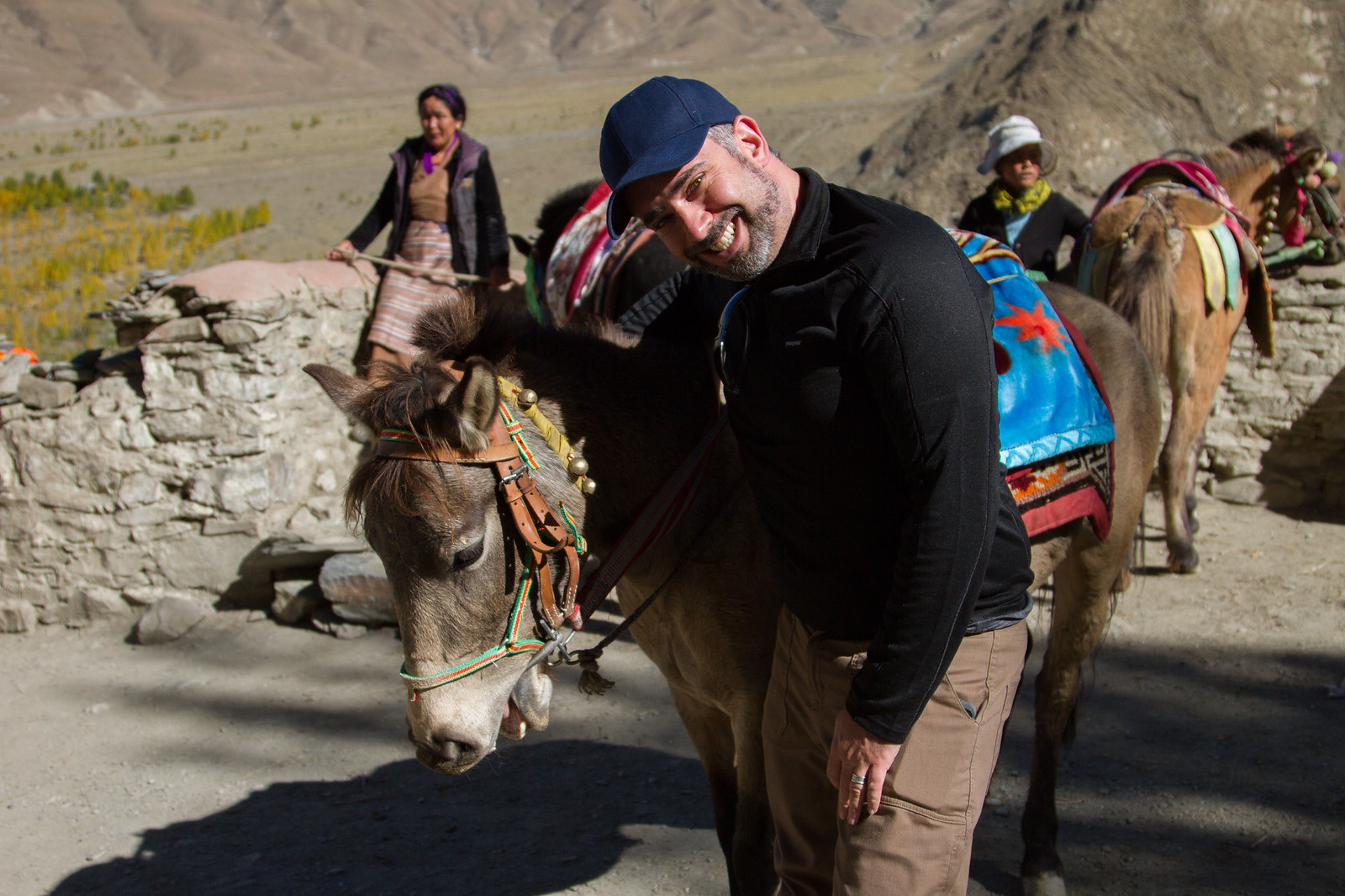 Smiling man in a blue cap leans beside a bridled pony as Tibetan women and pack ponies wait on a dusty mountain trail.