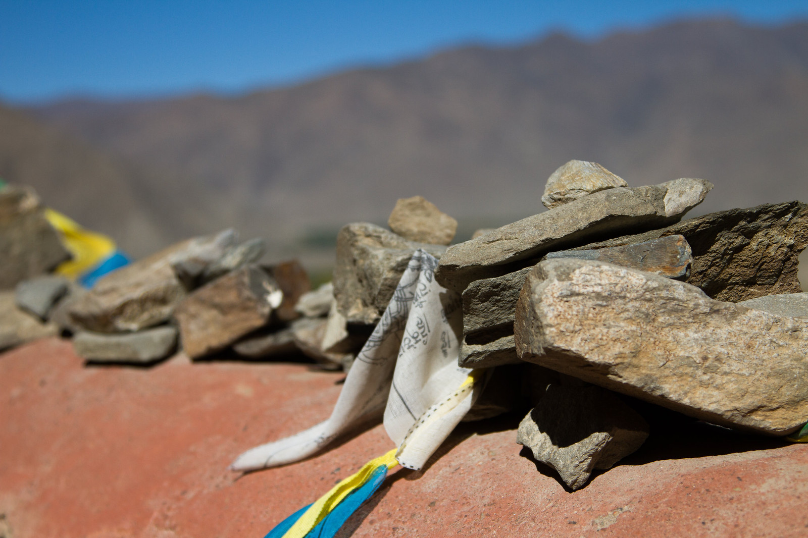 Stacked stones pinning white and colored Tibetan prayer flags against a backdrop of arid mountains.
