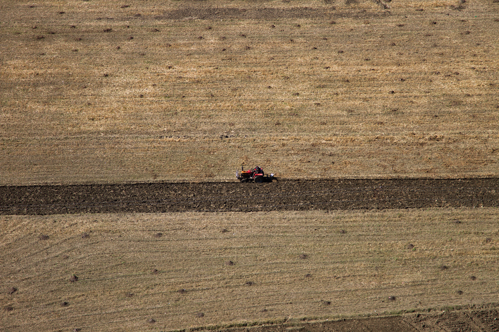 A lone red tractor plows a dark furrow across a vast expanse of dry, tan farmland.