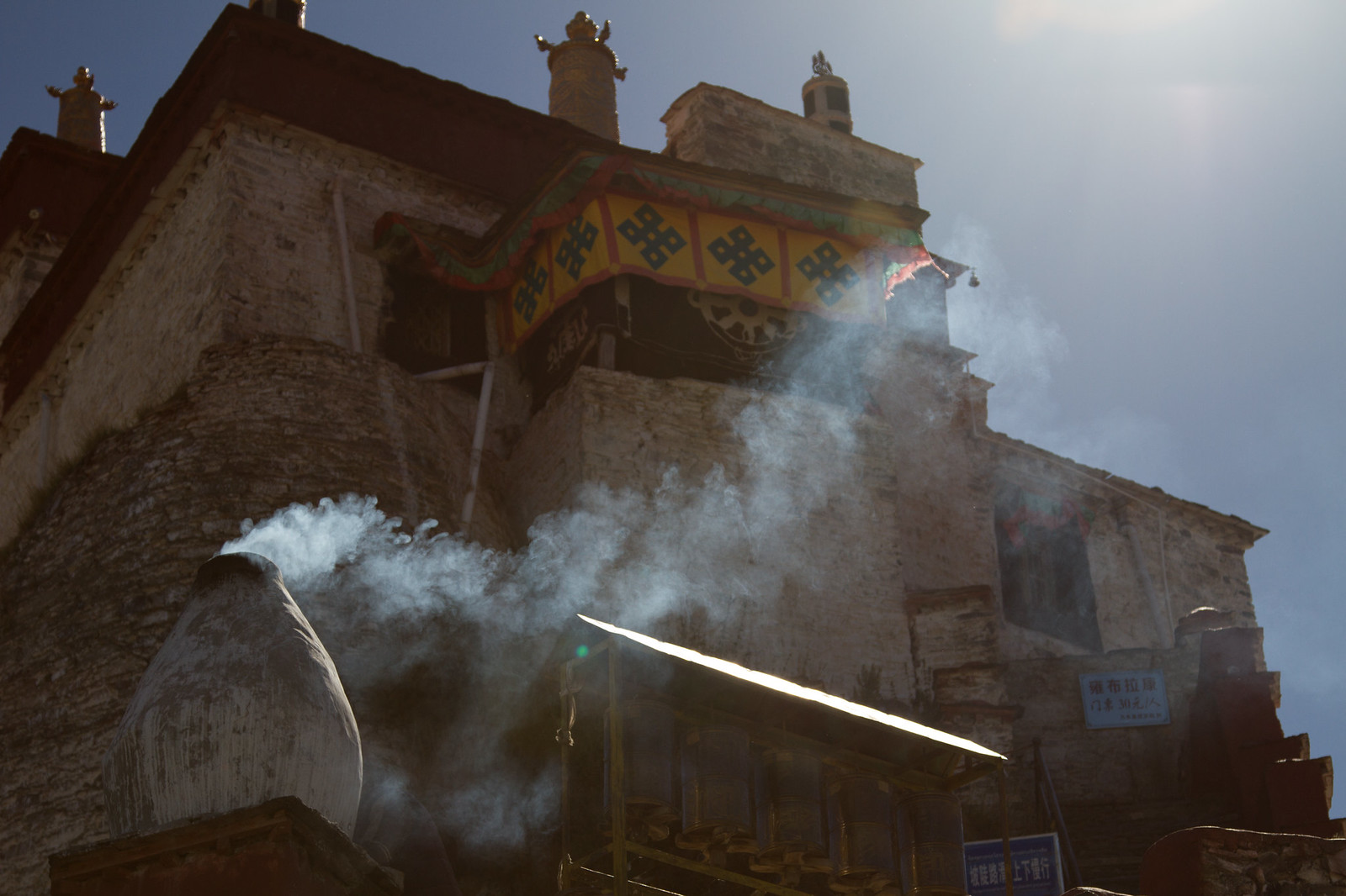 Incense smoke drifts from a whitewashed burner before a sunlit Tibetan monastery with an ornate gold-and-blue eaved roof.