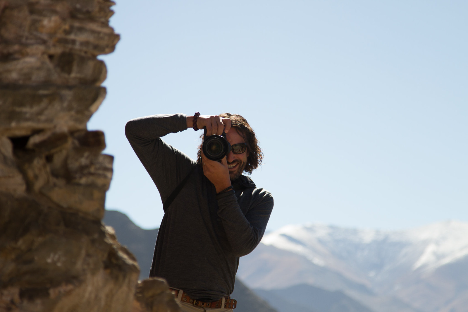 Smiling man in sunglasses aims a DSLR camera beside a rock outcrop, snow-capped mountains behind him.