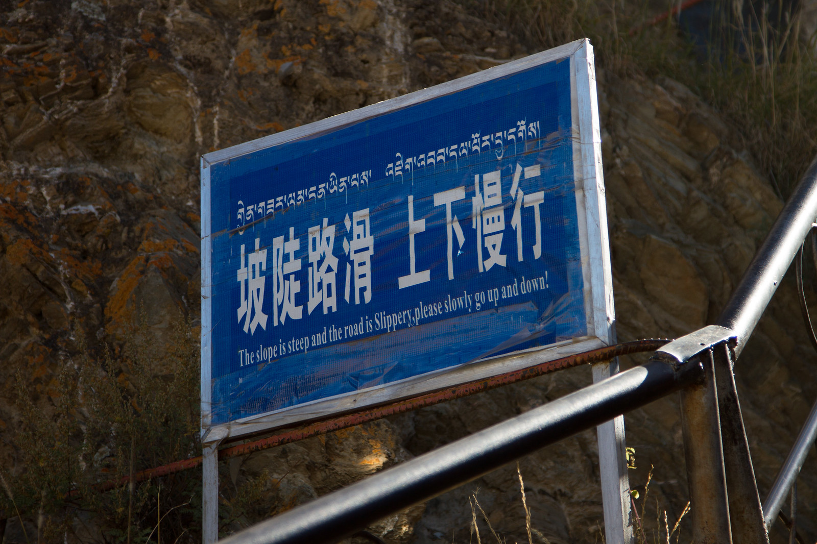 Blue roadside sign in Tibetan and Chinese reading "The slope is steep and the road is Slippery," against a rocky slope.