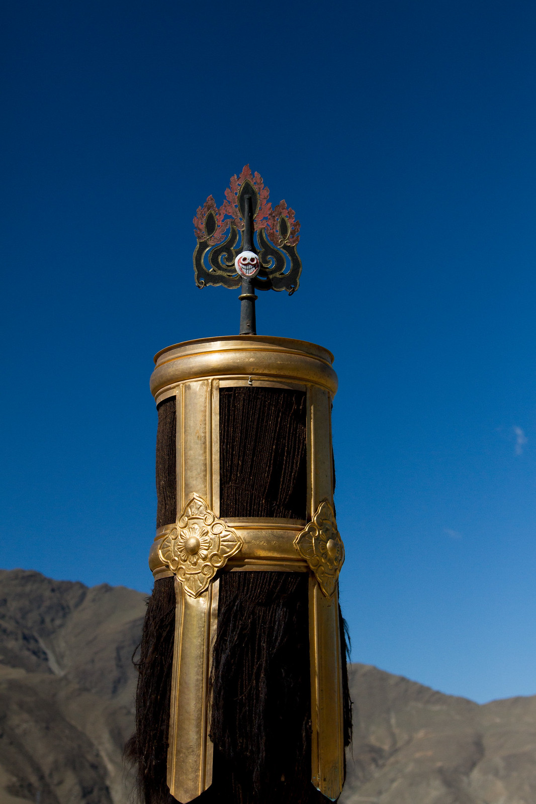 A gilded Tibetan rooftop banner ornament topped with a flame crest against a deep blue sky and barren mountains.