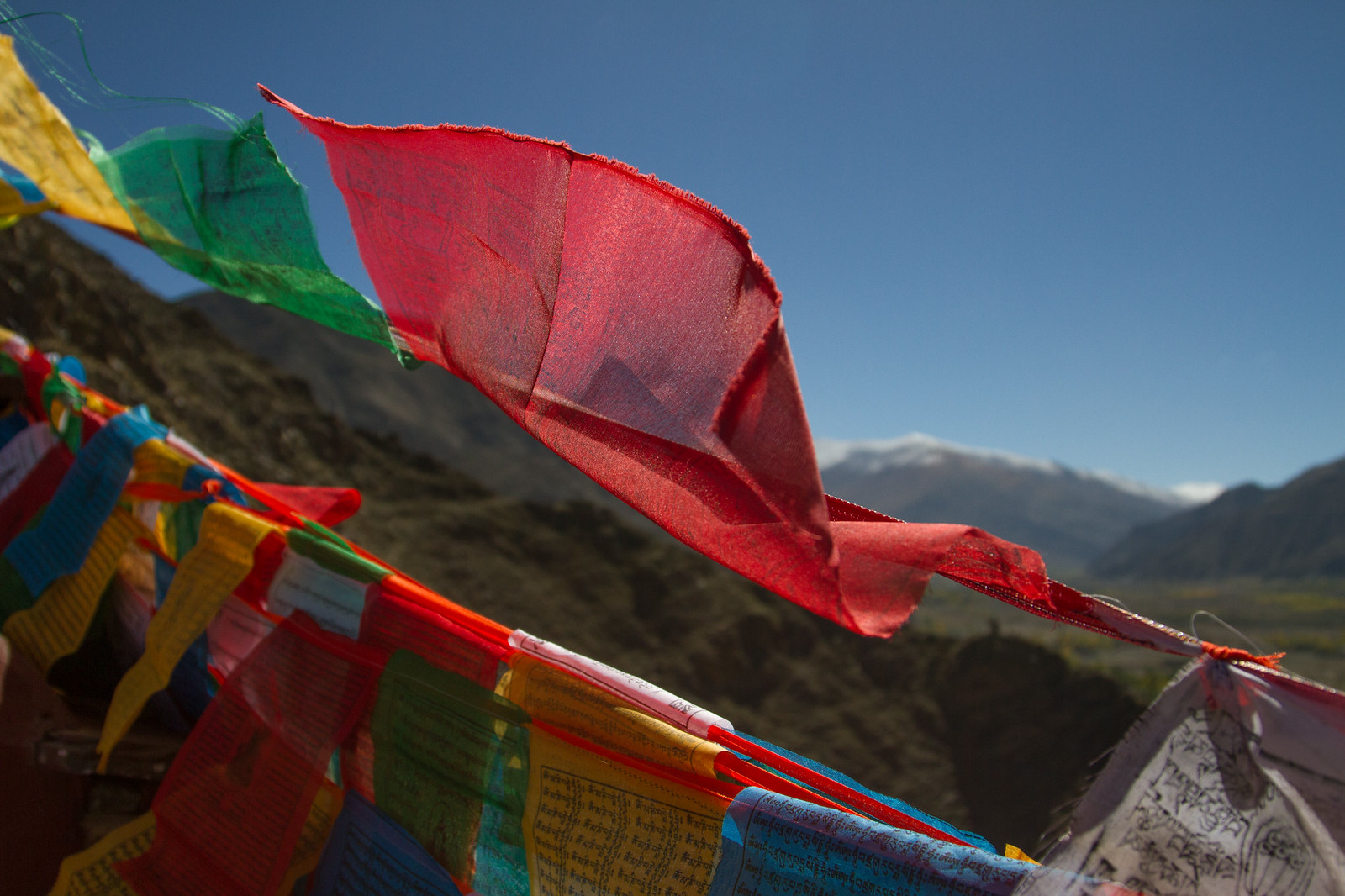 Colorful Tibetan prayer flags fluttering against a blue sky above snow-capped Himalayan mountains and arid valleys.