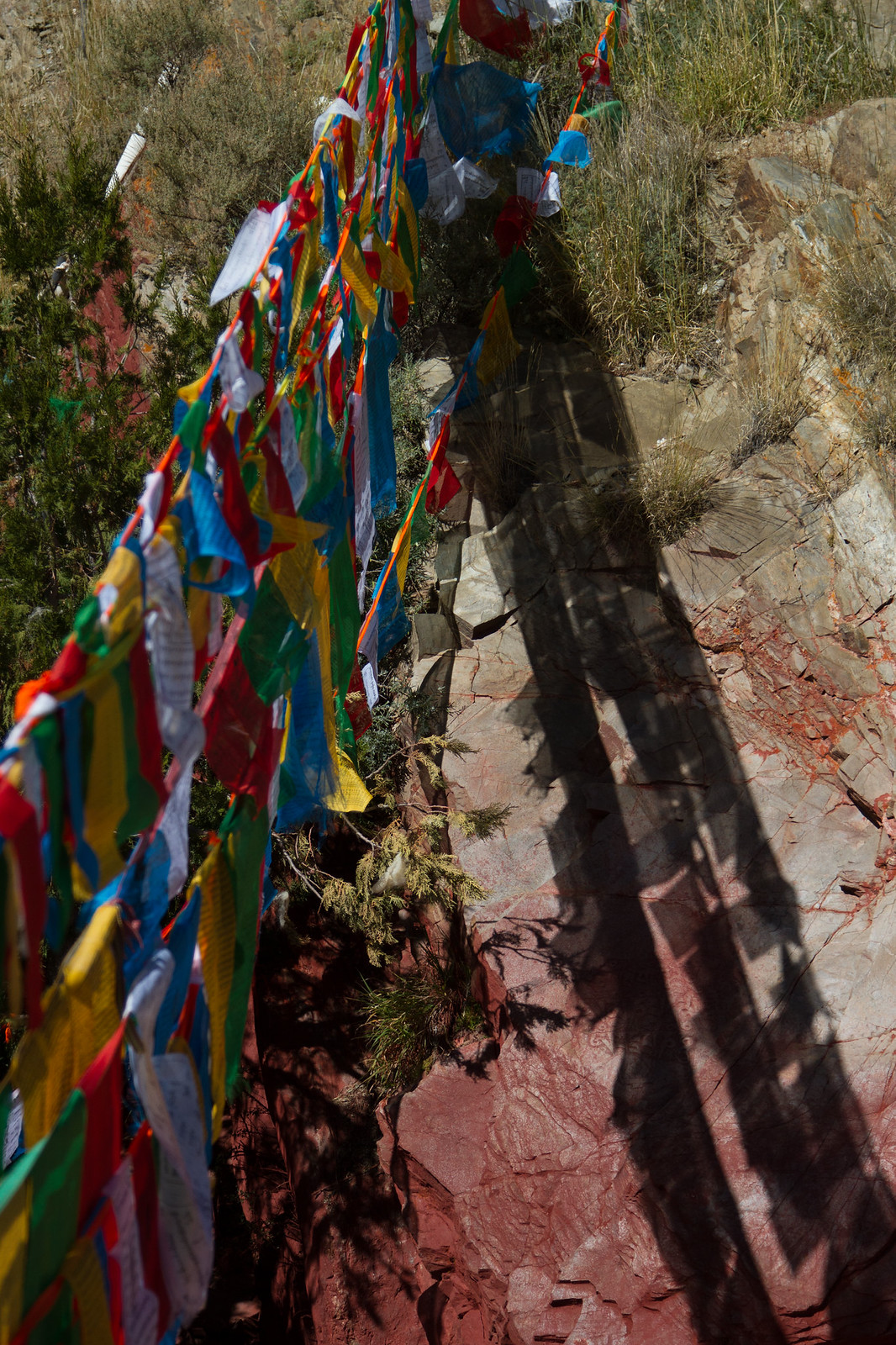 Colorful Tibetan prayer flags strung along a rocky red hillside trail in bright sunlight.
