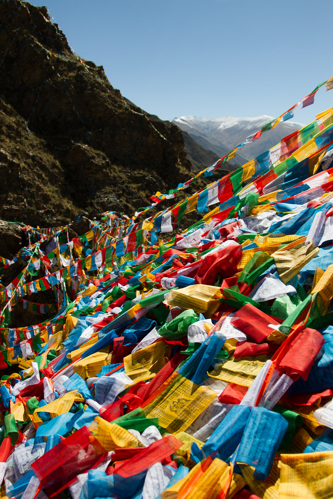Masses of colorful Tibetan prayer flags strung across a mountain pass with snow-capped peaks beyond.