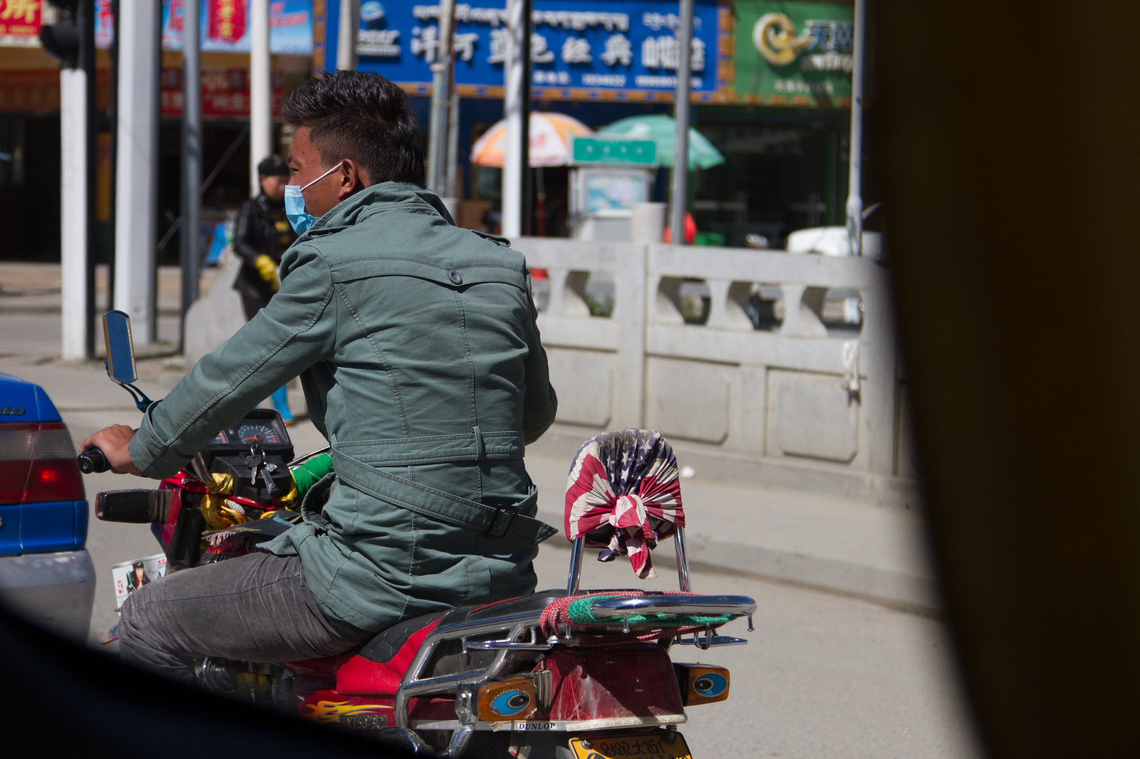 Man in a green jacket and face mask on a motorbike with a US-flag-covered backrest, on a Tibetan street.