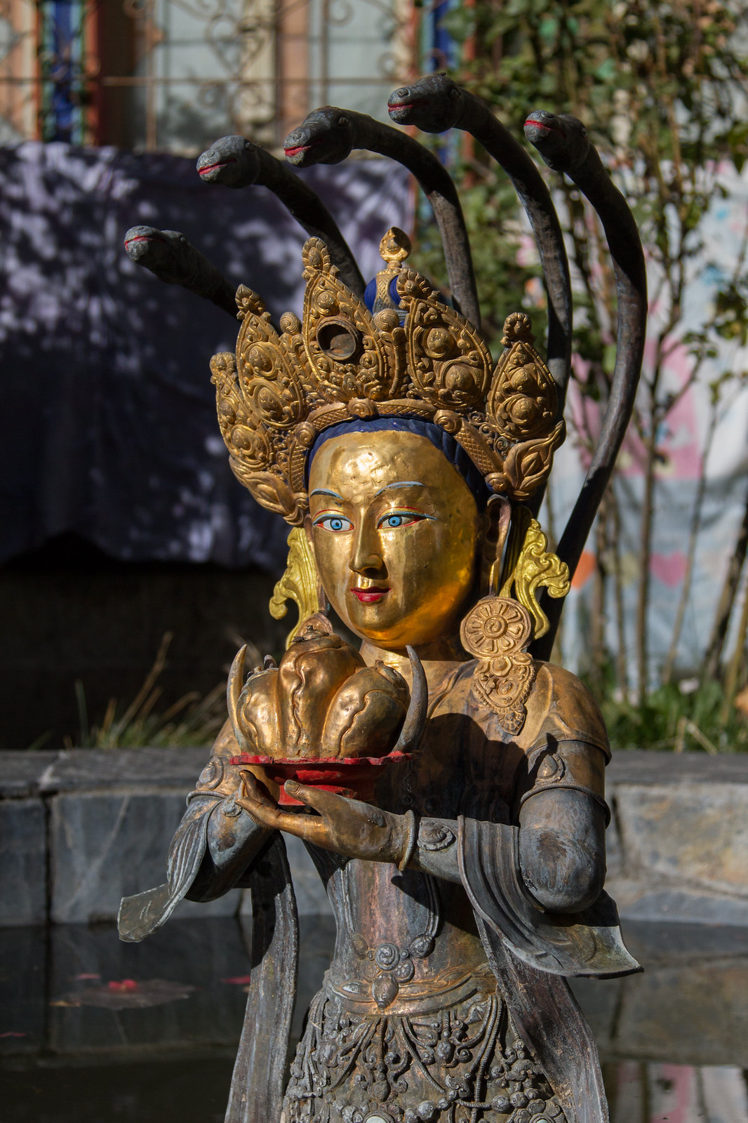 Gilded Tibetan deity statue with a jeweled crown and cobra hoods, holding an offering bowl.
