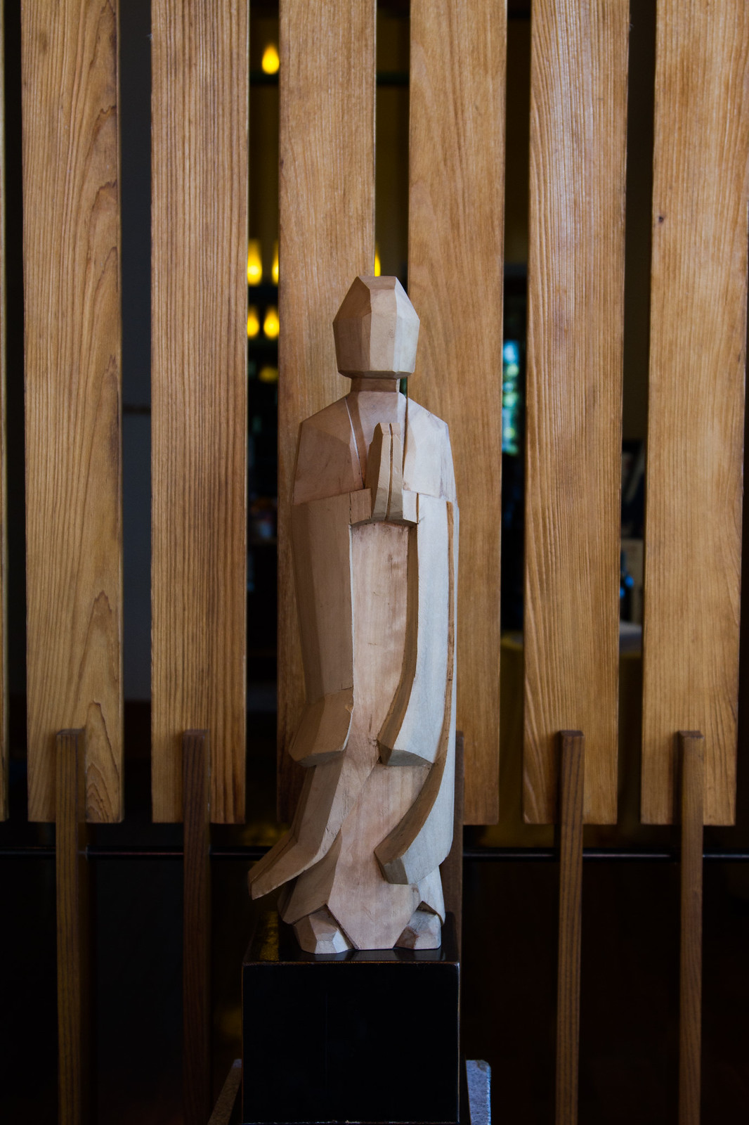 Angular carved wooden statue of a robed figure displayed on a pedestal before a wooden slat screen.