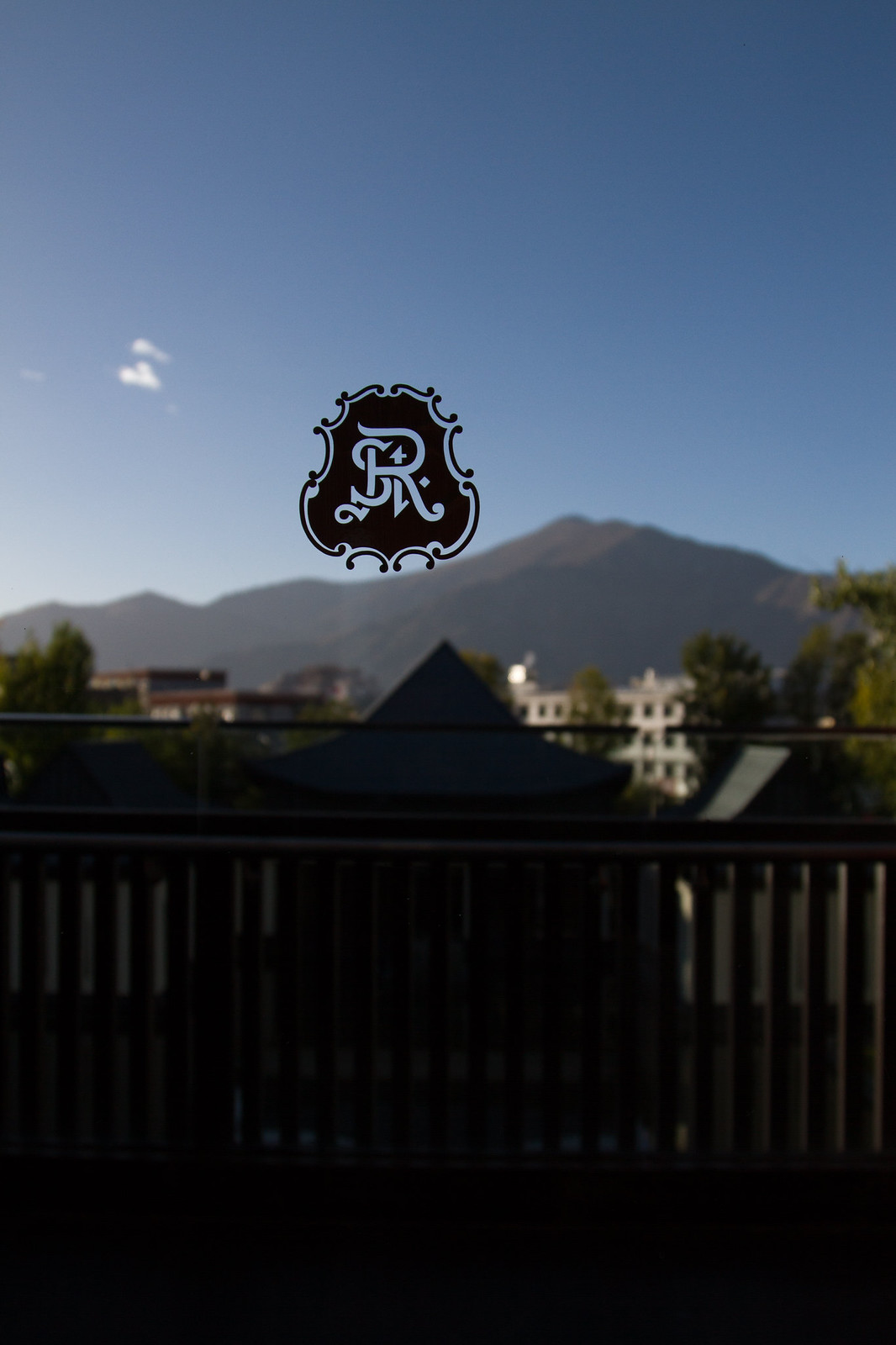 An ornate "R" crest on a window overlooking mountains and rooftops in Lhasa under a clear blue sky.