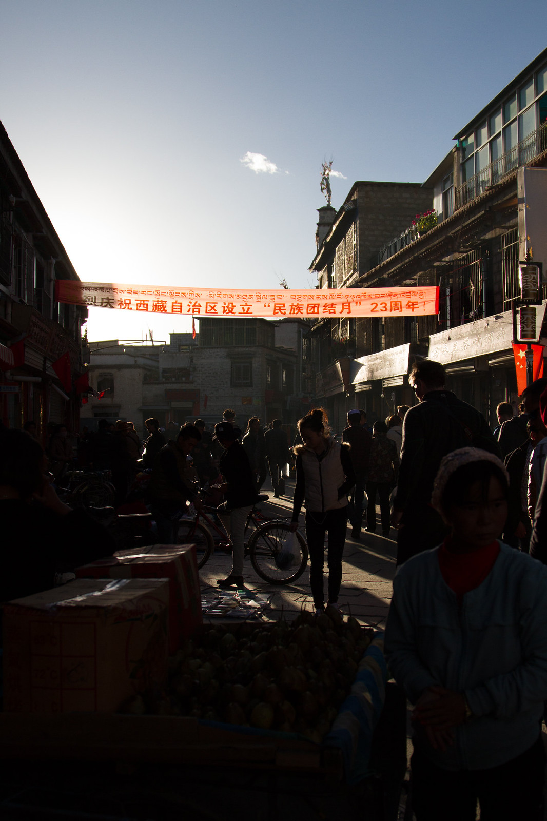Backlit Tibetan market street at dusk with crowds beneath a red banner reading "庆祝西藏自治区设立23周年