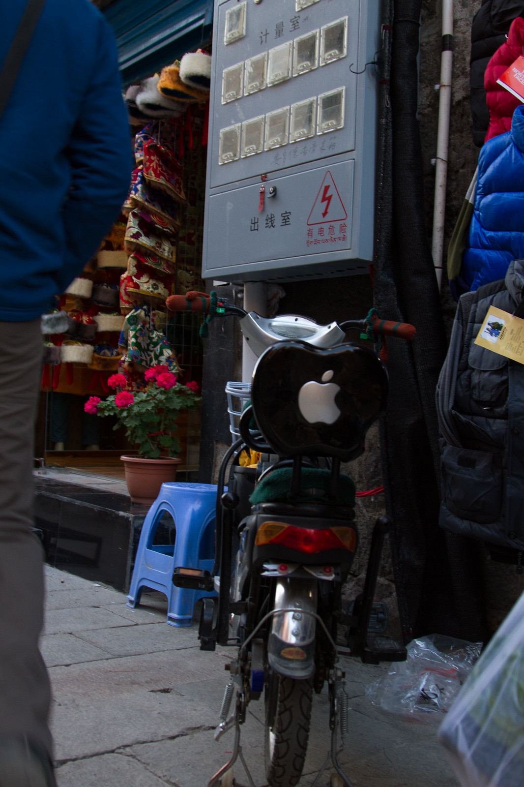 Electric bike with an Apple logo backrest parked by a Tibetan shop selling fur hats, beside a power box.